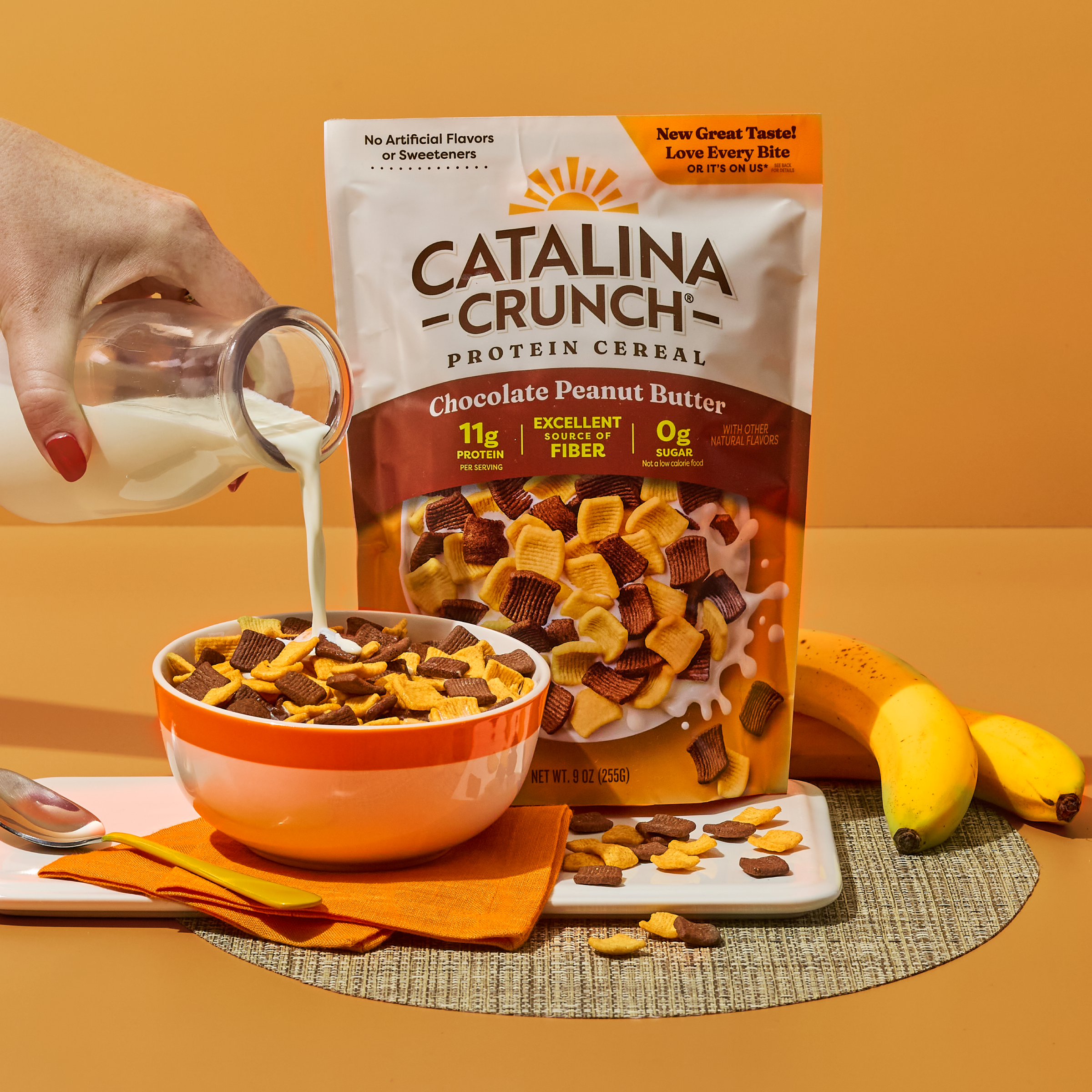 Catalina Crunch Protein Cereal being poured into a bowl with milk, surrounded by bananas on an orange background.