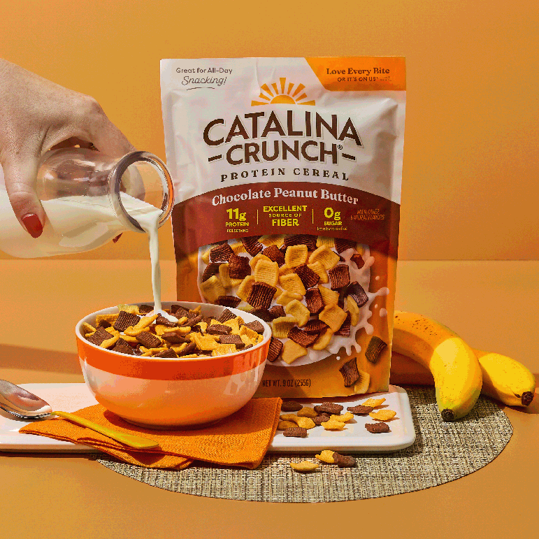 Catalina Crunch Protein Cereal being poured into a bowl with milk, surrounded by bananas on an orange background.