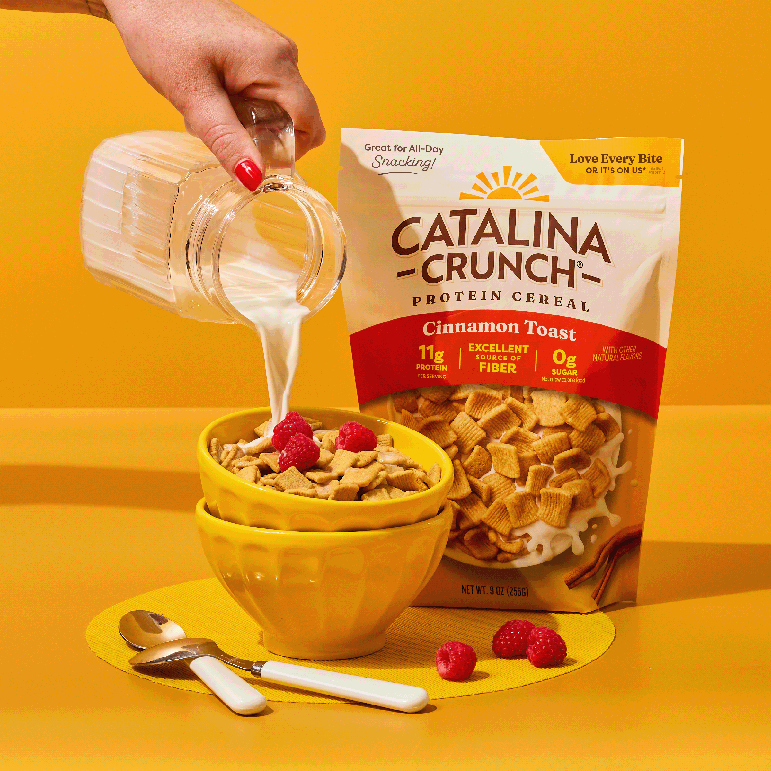 Catalina Crunch Protein Cereal being poured into a bowl with raspberries on a yellow background