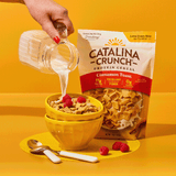 Catalina Crunch Protein Cereal being poured into a bowl with raspberries on a yellow background