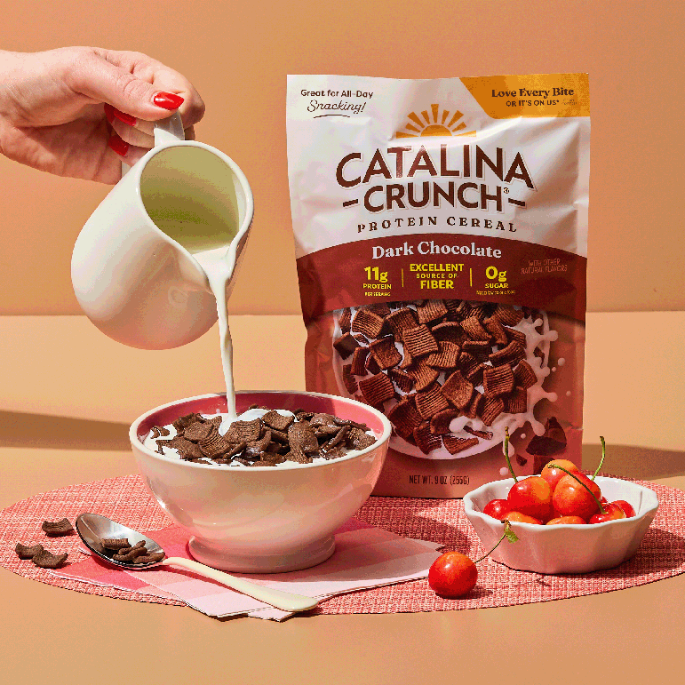 Catalina Crunch protein cereal being poured into a bowl with a pitcher of milk on a peach background.