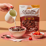 Catalina Crunch protein cereal being poured into a bowl with a pitcher of milk on a peach background.