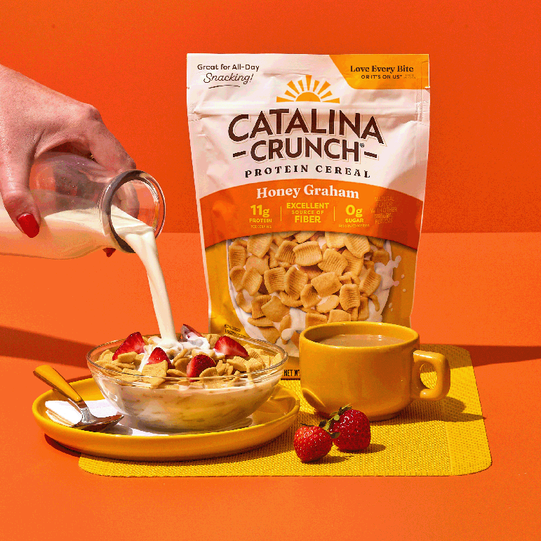 Catalina Crunch Protein Cereal being poured into a bowl with strawberries and a cup of coffee on an orange background.