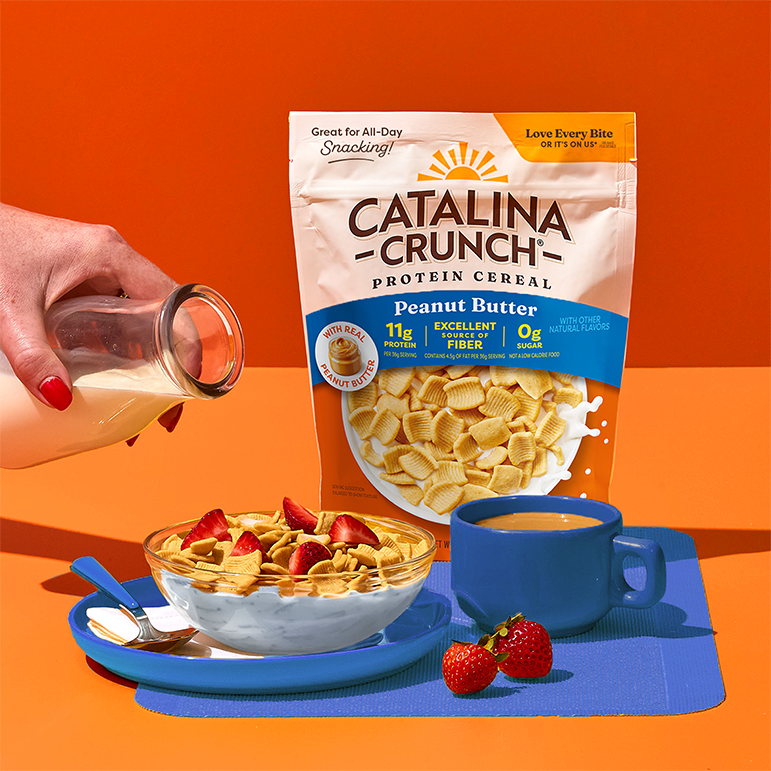 Catalina Crunch Peanut Butter protein cereal with a bowl of cereal and strawberries and a cup of coffee on an orange background.