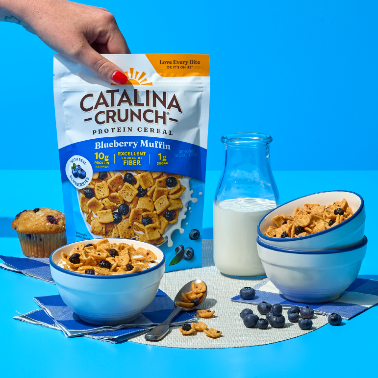 Catalina Crunch protein cereal with bowls, milk, and blueberries on a blue background